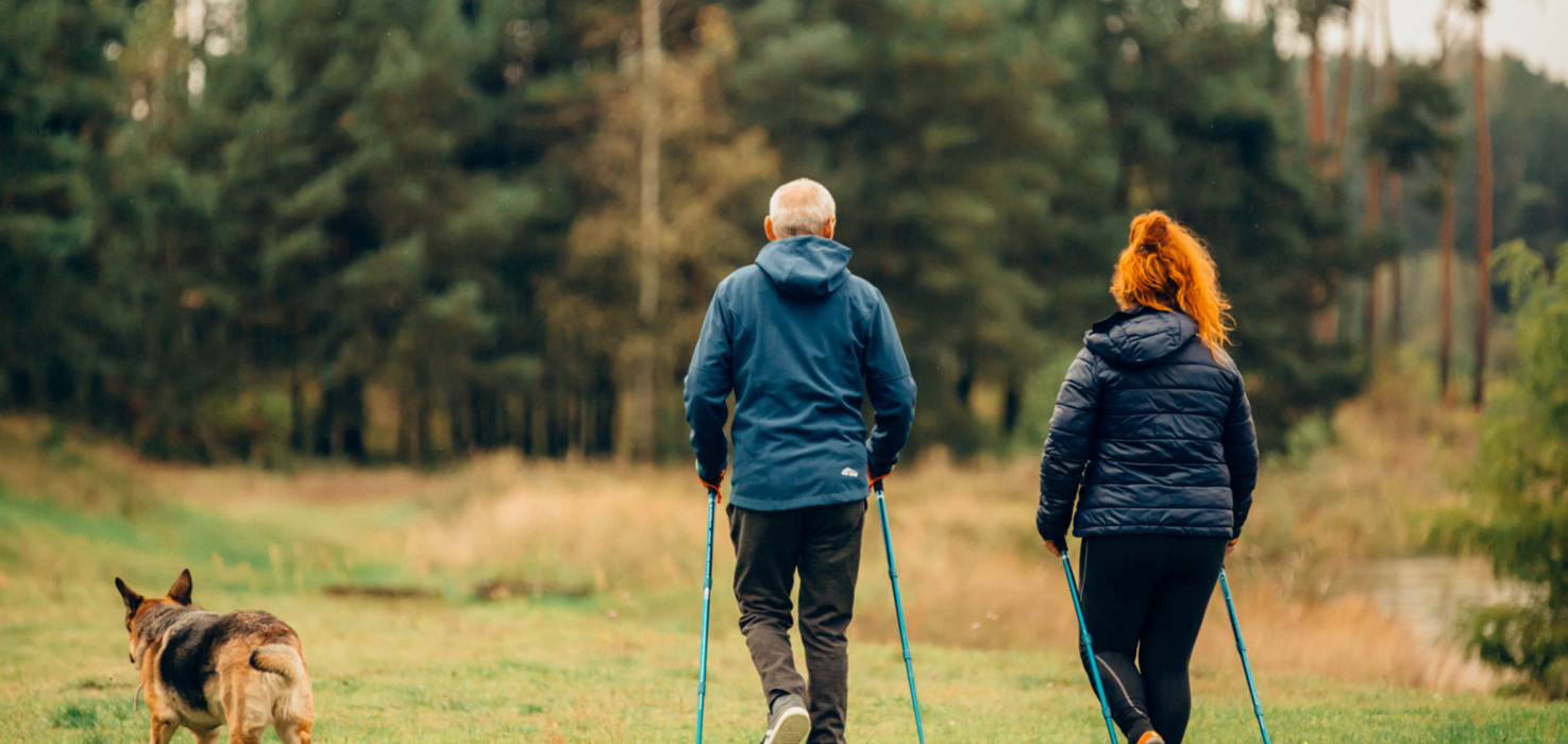 Para seniorów w jesiennej odzieży spacerująca z kijkami po polanie z psem, otoczeni drzewami i zielenią.