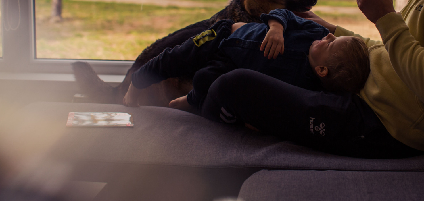 Dziecko leżące na kolanach dorosłego z filiżanką, obok pies, odpoczywający na kanapie przy oknie.