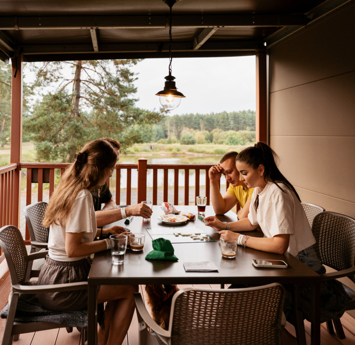 Grupa gości grających w planszówkę na tarasie domku mobilnego z widokiem na staw i rzekę Wartę.