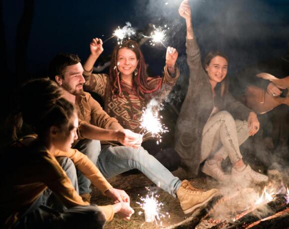 Grupa przyjaciół siedząca wieczorem przy ognisku, trzymająca zimne ognie i grająca na gitarze.