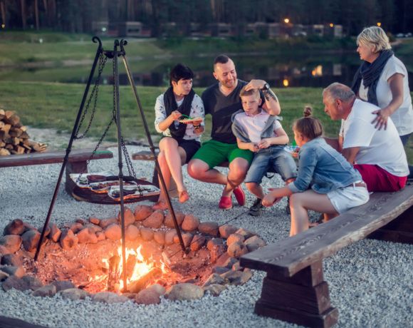 Rodzina relaksująca się przy ognisku z rusztem do grillowania, siedząca na drewnianych ławkach nad rzeką Wartą o zmierzchu.