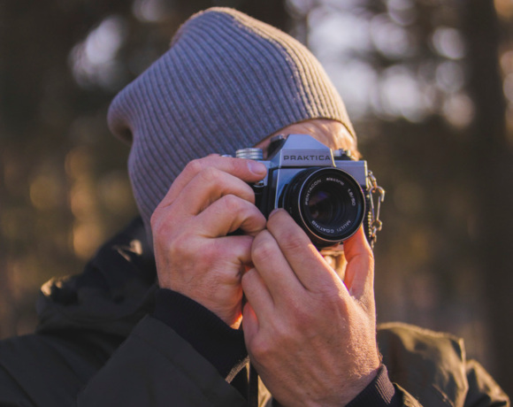 Mężczyzna w ciepłej czapce robiąca zdjęcie aparatem fotograficznym na tle jesiennego lasu.