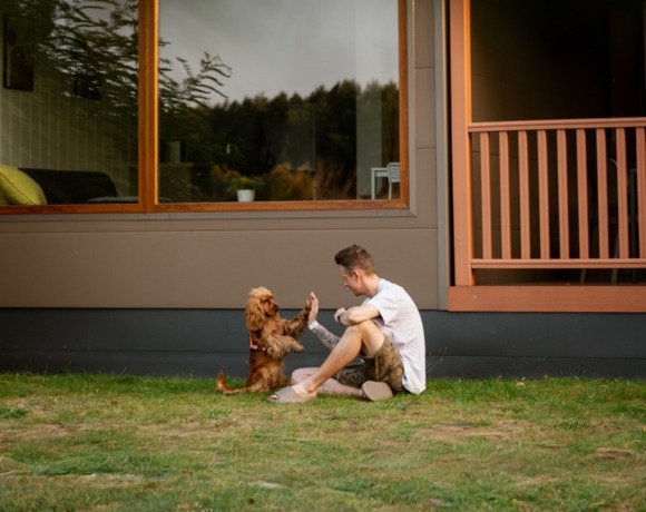 Nowoczesny domek mobilny z widokiem na staw, dużym oknem i tarasem, mężczyzna bawi się z małym psem na trawie.