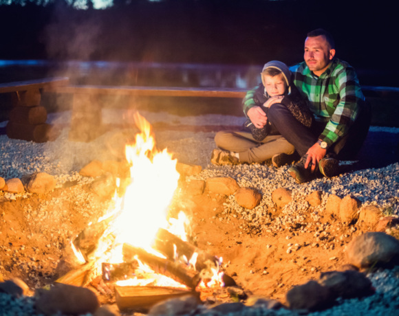 Mężczyzna i dziecko ogrzewają się przy ognisku na zewnątrz, siedząc na kamienistym podłożu nocą.