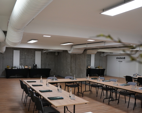 Nowoczesna sala konferencyjna z układem stołów w kształcie litery U, wodą i notatnikami oraz surowymi betonowymi ścianami.