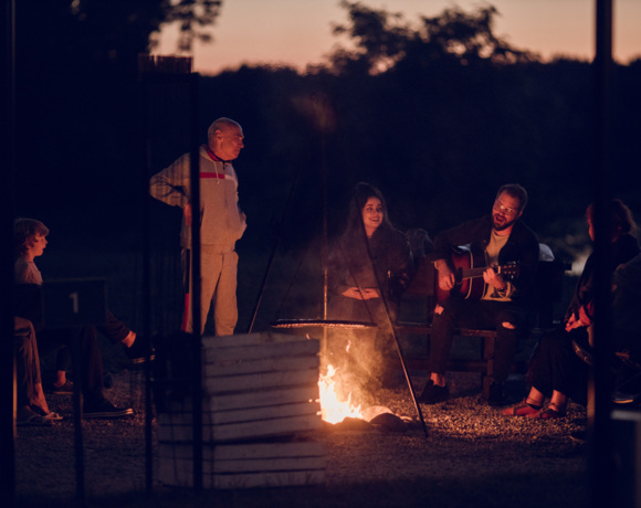 Goście Resortu Stara Wieś siedzący wieczorem wokół ogniska, z jedną osobą grającą na gitarze, tworzący przyjazną atmosferę.