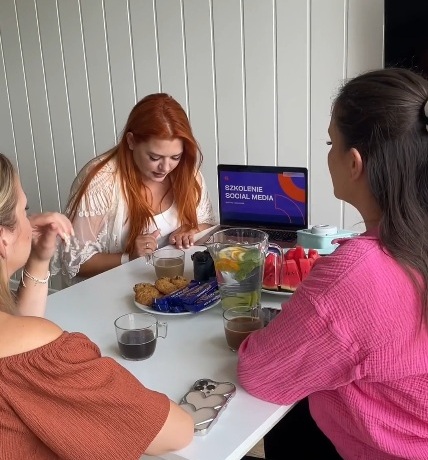 Trzy kobiety siedzące przy stole z przekąskami i napojami, uczestniczące w szkoleniu z social mediów na laptopie.