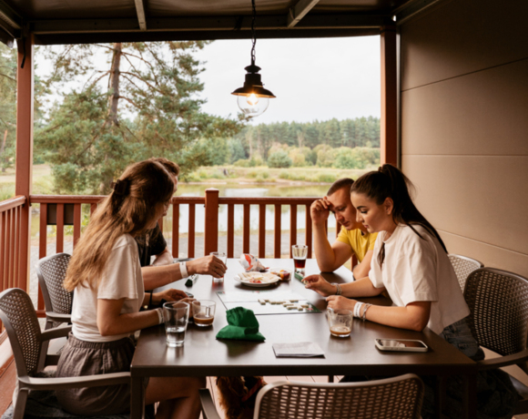 Grupa gości grających w planszówkę na tarasie domku mobilnego z widokiem na staw i rzekę Wartę.