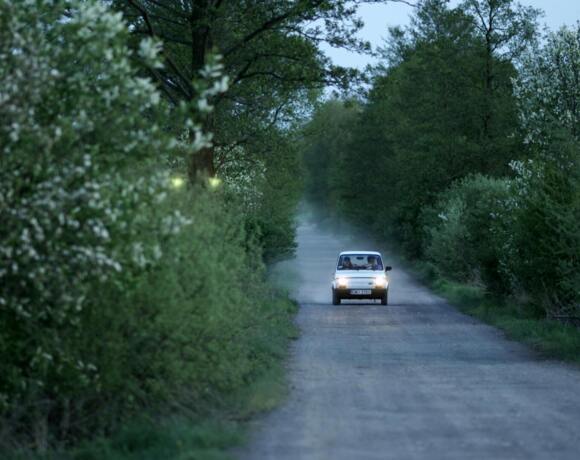 Biały samochód jedzie podmiejską, leśną drogą otoczoną zielonymi drzewami i krzewami o zmierzchu.