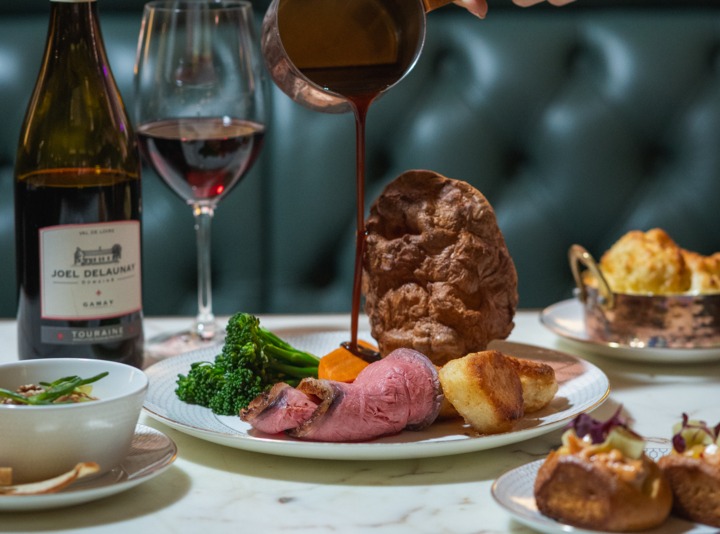 Hand pouring gravy over roast beef, Yorkshire pudding and roast potatoes on a plated Sunday roast with red wine.
