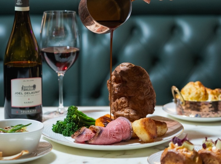 Roast dinner: sliced beef, Yorkshire pudding and roast potatoes; gravy poured from copper jug, red wine bottle and glass.