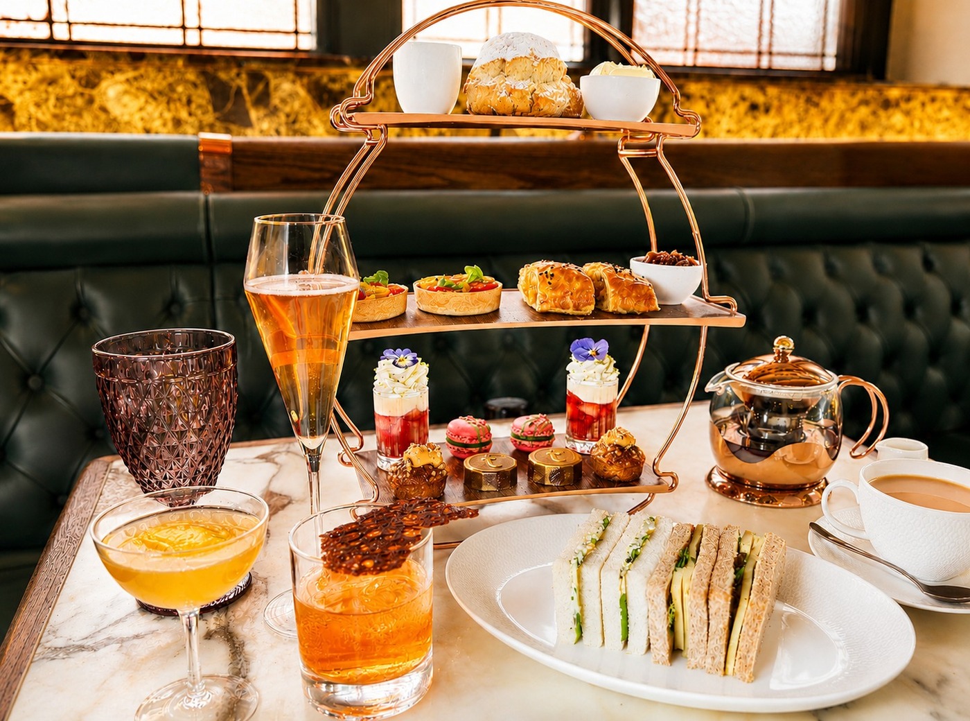 Three-tier afternoon tea stand with assorted pastries, sandwiches, cocktails and teapot on marble table in hotel lounge.