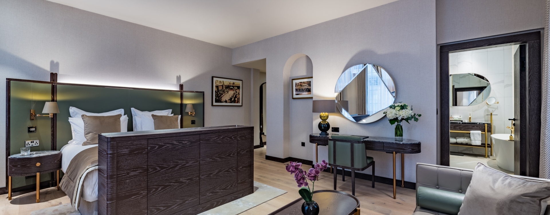Spacious suite with king bed, green headboard, seating area, desk with round mirror, flowers and marble bathroom with freestanding tub.