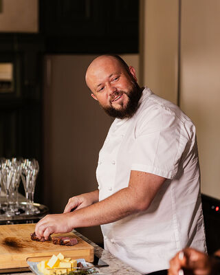 Chef in white jacket slicing roasted meat on wooden board at hotel restaurant counter with butter, herbs and wine glasses.