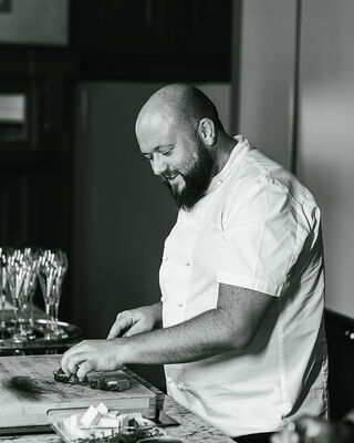 Hotel chef in white jacket slicing steak on a wooden board at a kitchen counter, preparing plates with glassware nearby