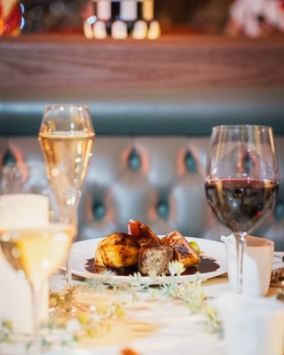Plated roasted meat with glossy jus, champagne flute and red wine glass on a white-linen table in an intimate tufted-banquette restaurant