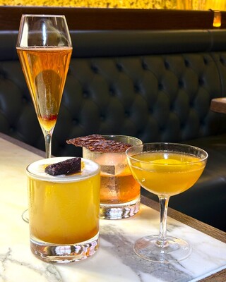 Four cocktails on a marble table before a tufted leather banquette: sparkling flute, foamy citrus with garnish, amber rocks with tuile, yellow coupe.