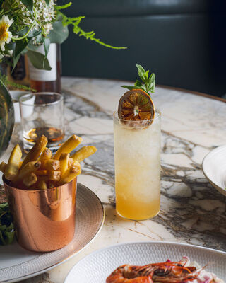 Tall citrus cocktail with crushed ice, dried orange and mint garnish beside copper cup of fries and prawn salad on marble table.