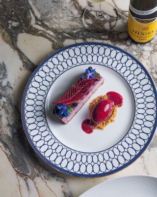 Plated berry dessert: purple mousse with red chocolate leaf, sorbet on crumble, edible flowers; wine bottle on marble.