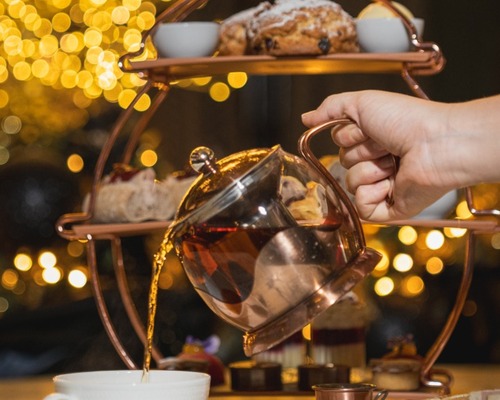 Hand pouring tea from copper pot into white cup; three-tier stand with scones, pastries and sandwiches, twinkling lights.