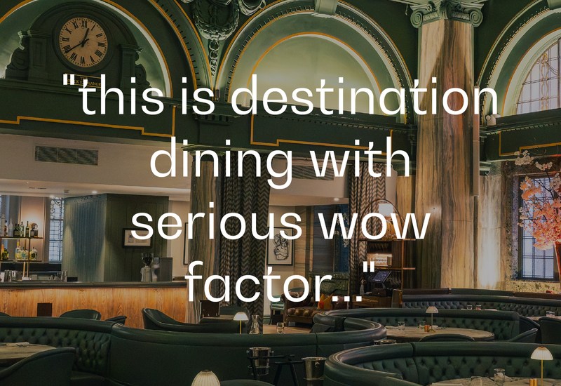 Grand hotel dining lounge with ornate arched ceiling and clock, marble columns, curved emerald banquettes, intimate tables and soft lamp lighting.