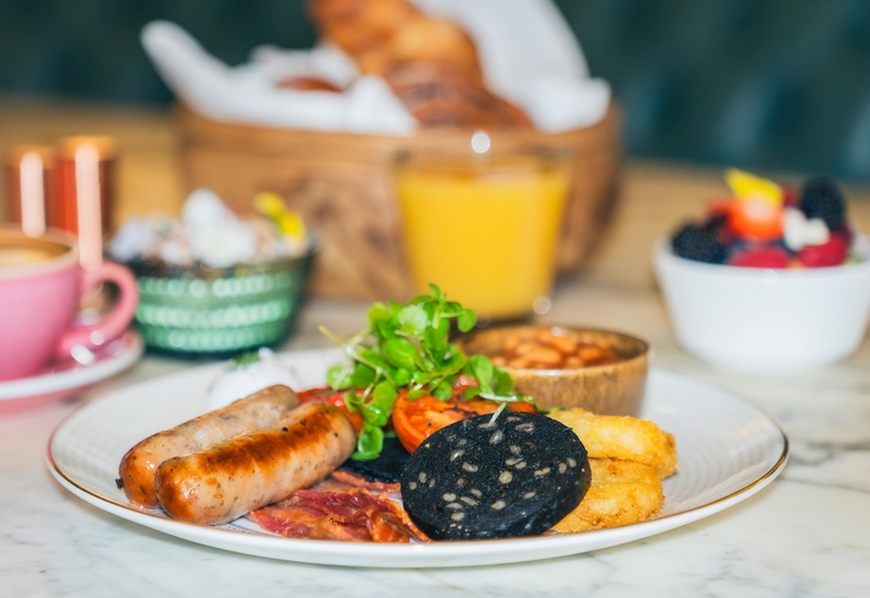 Full English breakfast: sausages, black pudding, bacon, hash browns and grilled tomato; orange juice and coffee behind.