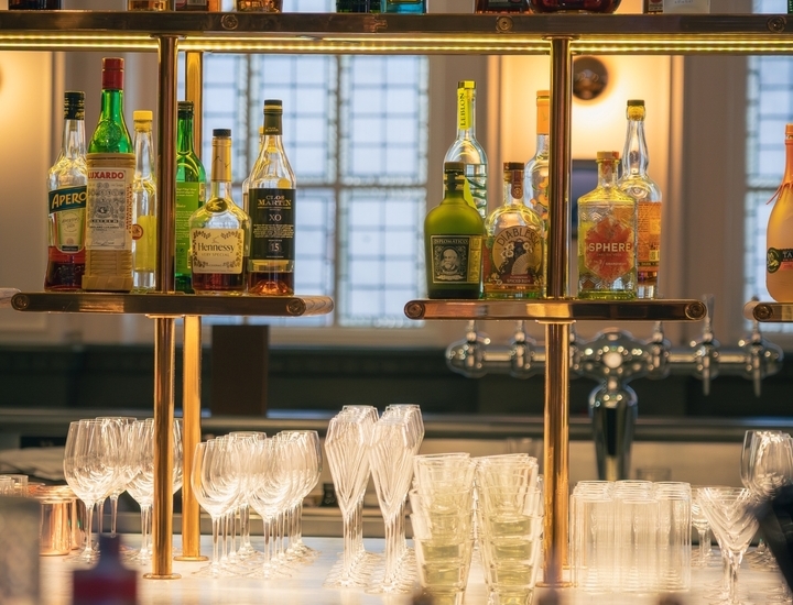Backlit hotel bar with premium spirits on brass shelves, stacked wine and cocktail glasses on marble counter, warm ambient light and leaded windows.