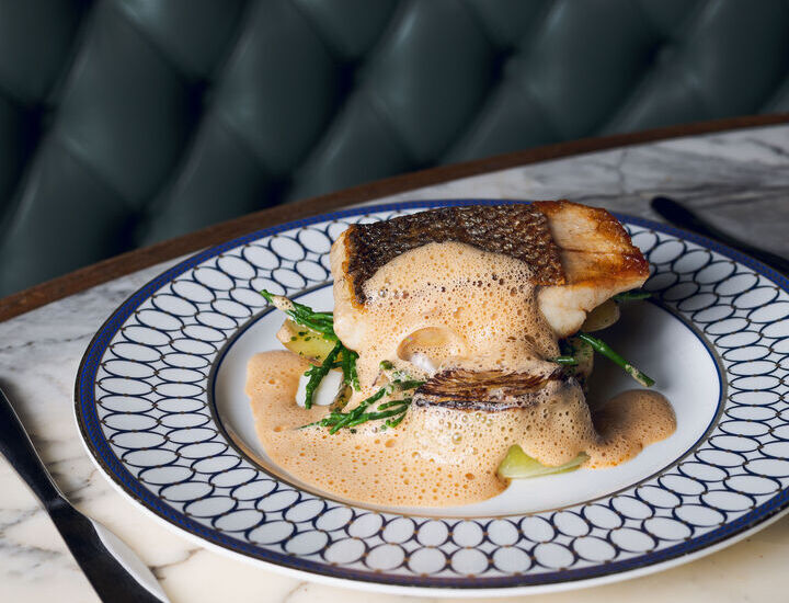 Pan-seared sea bass with frothy citrus sauce over greens, served on patterned china at marble table by tufted banquette.