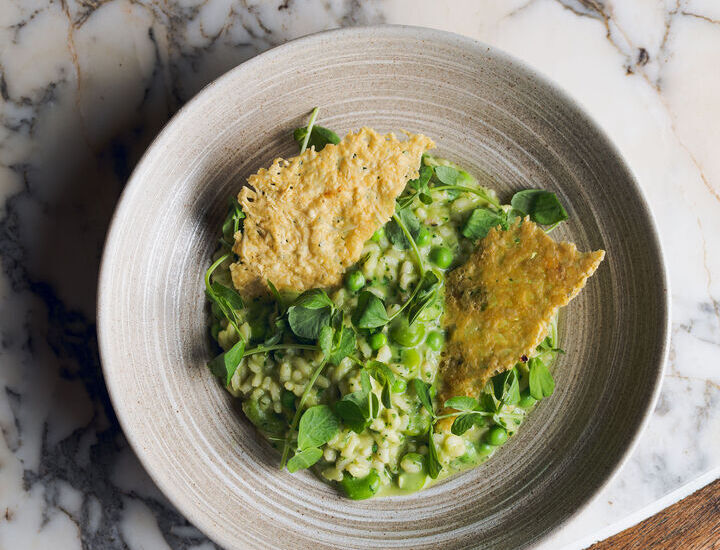 Creamy pea risotto with fresh peas, microgreens and two crisp Parmesan tuile in a rustic ceramic bowl on marble table.