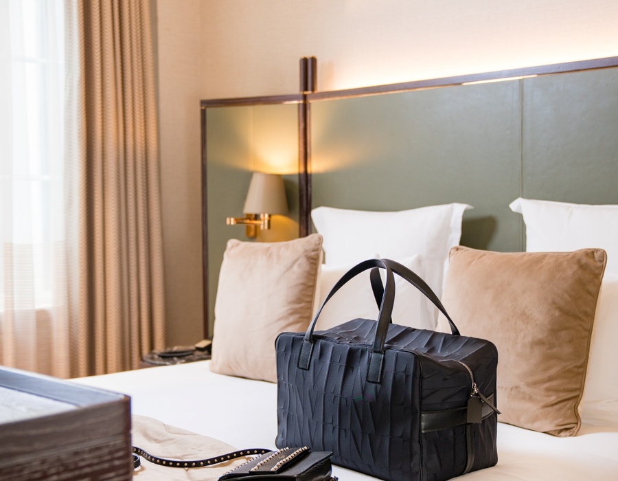 Plush king bed with padded green headboard, bedside lamp, beige pillows and a black travel bag on white linens.