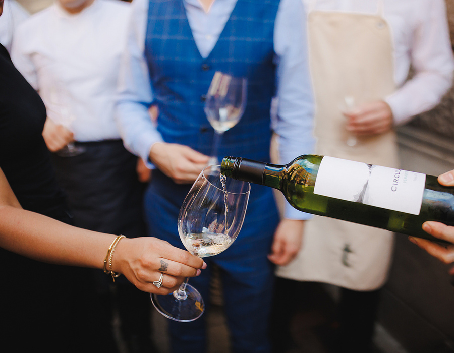 Server pouring white wine into guest's glass at a hotel wine-tasting reception, well-dressed guests and staff visible