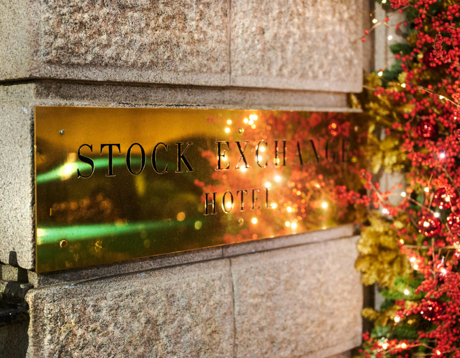 Brass plaque reading 'Stock Exchange Hotel' on stone wall, edged with red berry garland and twinkling string lights.