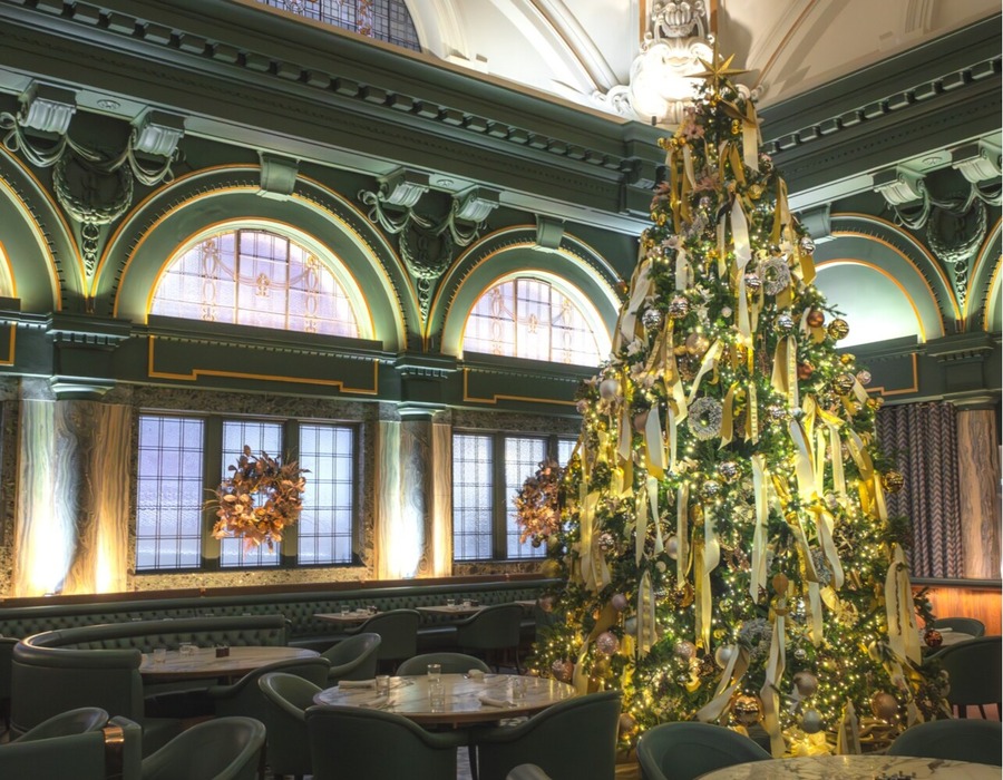 Hotel dining room with towering Christmas tree draped in gold ribbons and lights, arched windows and plush green seating.