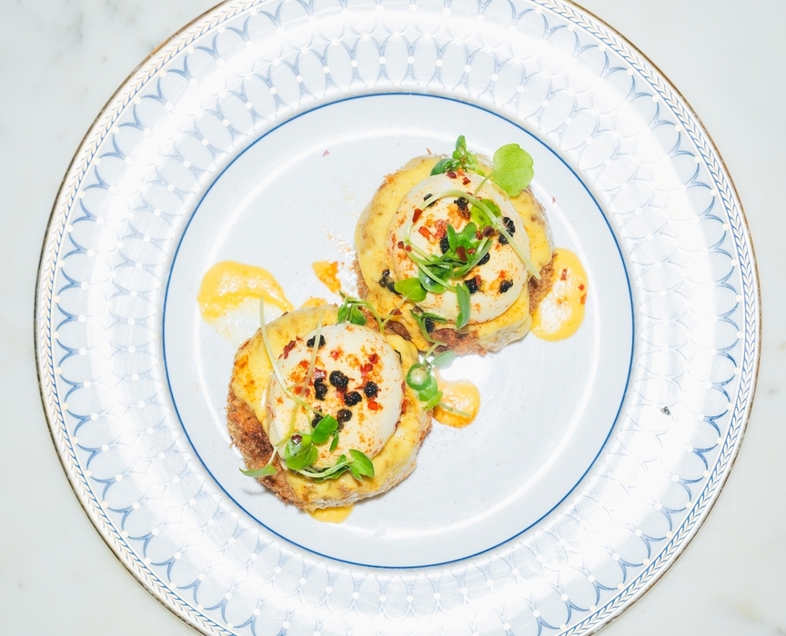 Two poached eggs with hollandaise and microgreens on toasted rounds, served on patterned porcelain plate over marble
