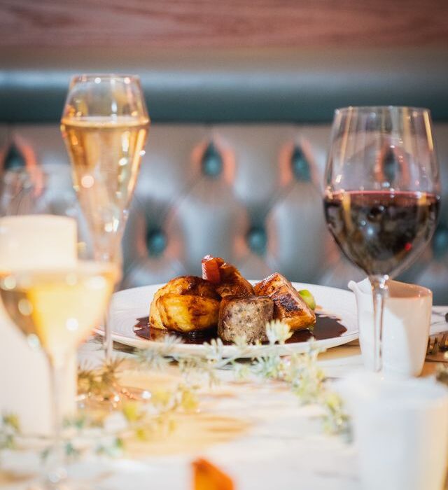 Plated roasted meat with glossy jus, champagne flute and red wine glass on a white-linen table in an intimate tufted-banquette restaurant