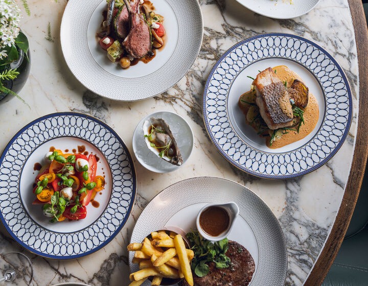 Elegant marble-top table set with chef-crafted dishes: lamb chops, seared fish, steak with fries and sauce, fresh salads, oyster and glass of red wine.