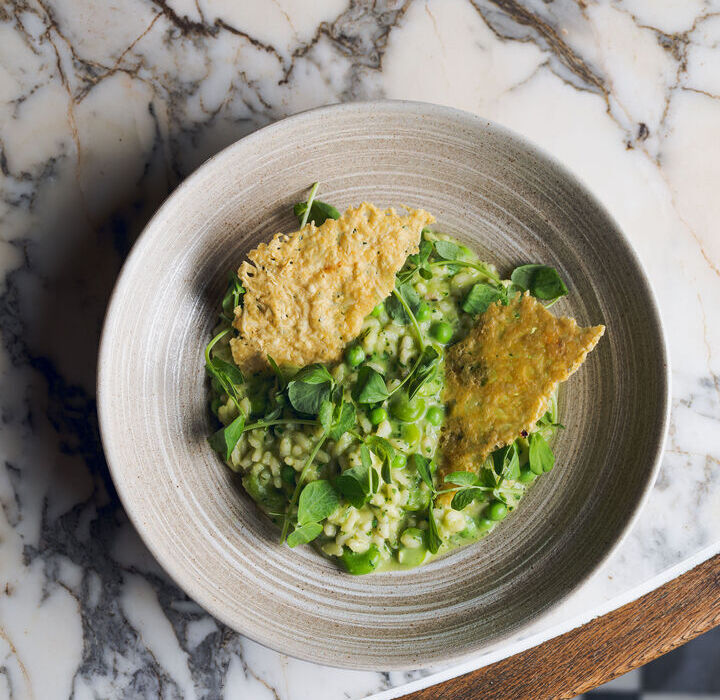 Creamy pea risotto with fresh peas, microgreens and two crisp Parmesan tuile in a rustic ceramic bowl on marble table.