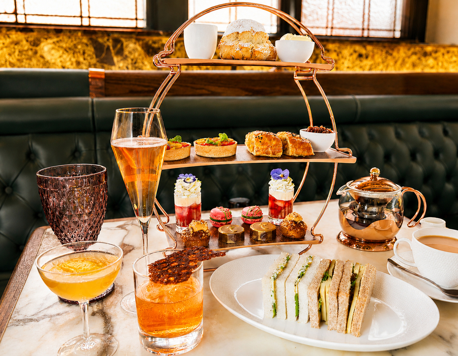 Three-tier afternoon tea stand with assorted pastries, sandwiches, cocktails and teapot on marble table in hotel lounge.
