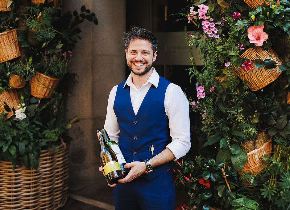 Smiling sommelier in blue vest holding two wine bottles at a restaurant doorway framed by wicker baskets and flowers