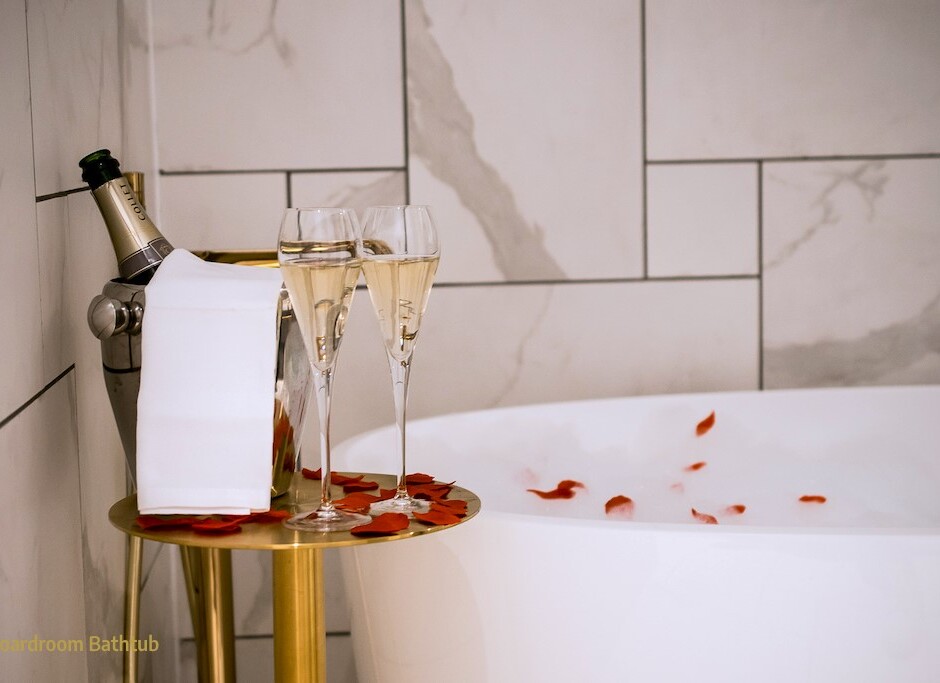 Freestanding bathtub with rose petals, marble tile wall, champagne in ice bucket and two flutes on a gold side table for two