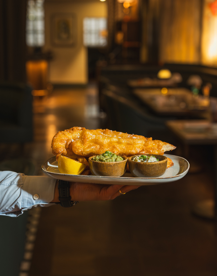 Waiter presenting a plate of golden beer-battered fish with lemon wedge and two dipping sauces in warm, cozy restaurant