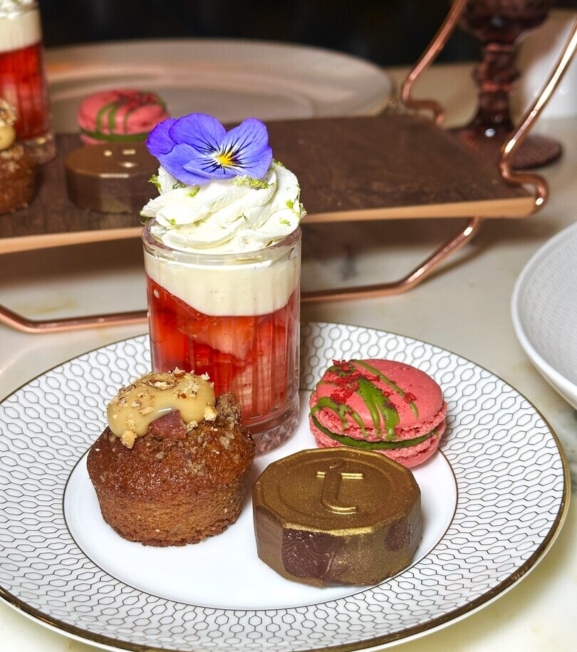 Assorted petit fours on china: strawberry parfait with cream and flower, pink macaron, caramel mini cake and gold chocolate.