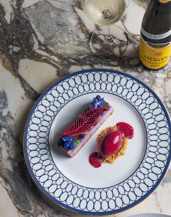 Plated berry dessert: purple mousse with red chocolate leaf, sorbet on crumble, edible flowers; wine bottle on marble.