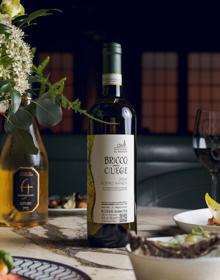 Bottled Italian Roero Arneis on marble table with floral arrangement, plated dishes and a wine glass in a refined restaurant.