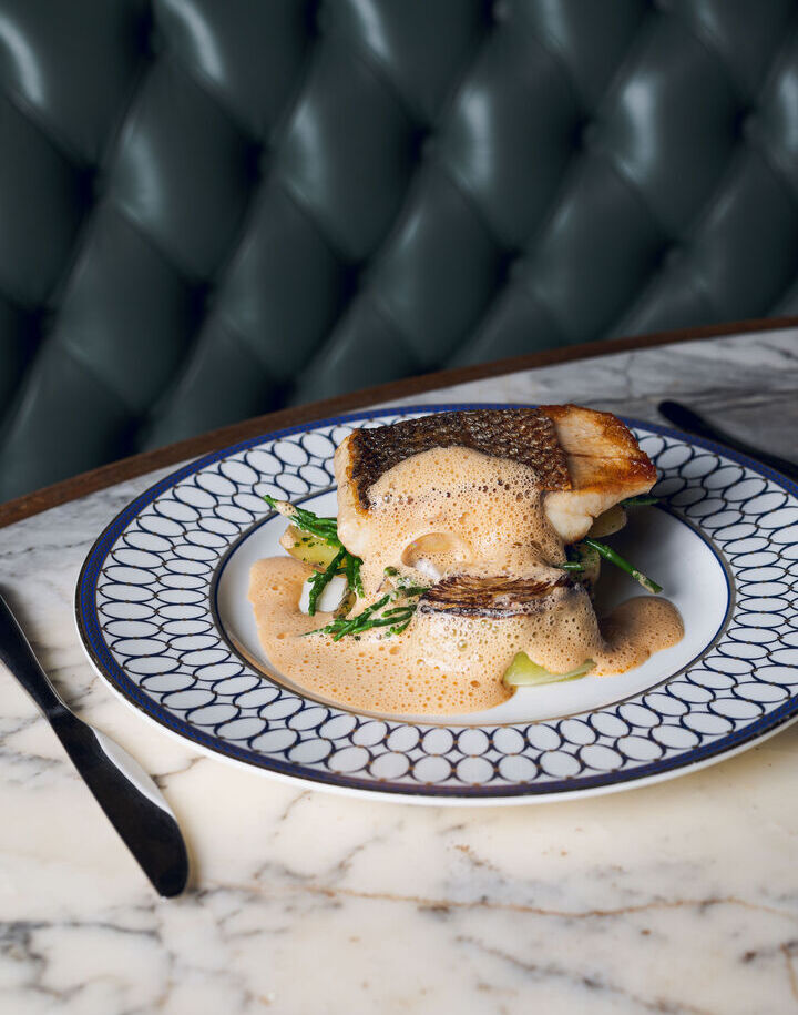 Pan-seared sea bass with frothy citrus sauce over greens, served on patterned china at marble table by tufted banquette.