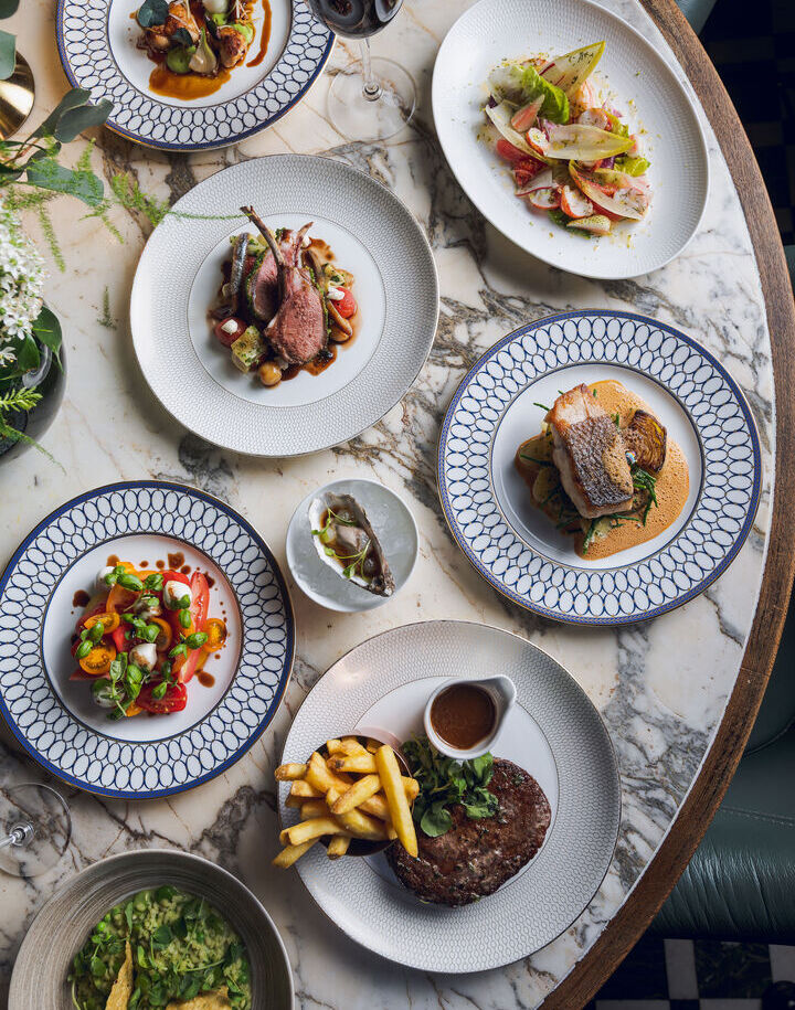 Elegant marble-top table set with chef-crafted dishes: lamb chops, seared fish, steak with fries and sauce, fresh salads, oyster and glass of red wine.