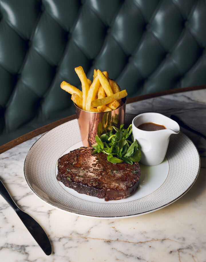 Grilled steak with watercress, fries in a copper cup and jus in a white pitcher on a marble table by a green tufted banquette.