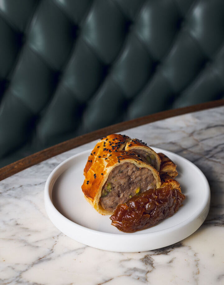 Flaky puff pastry sausage roll with pistachio, black sesame and onion chutney on marble table, tufted green banquette.
