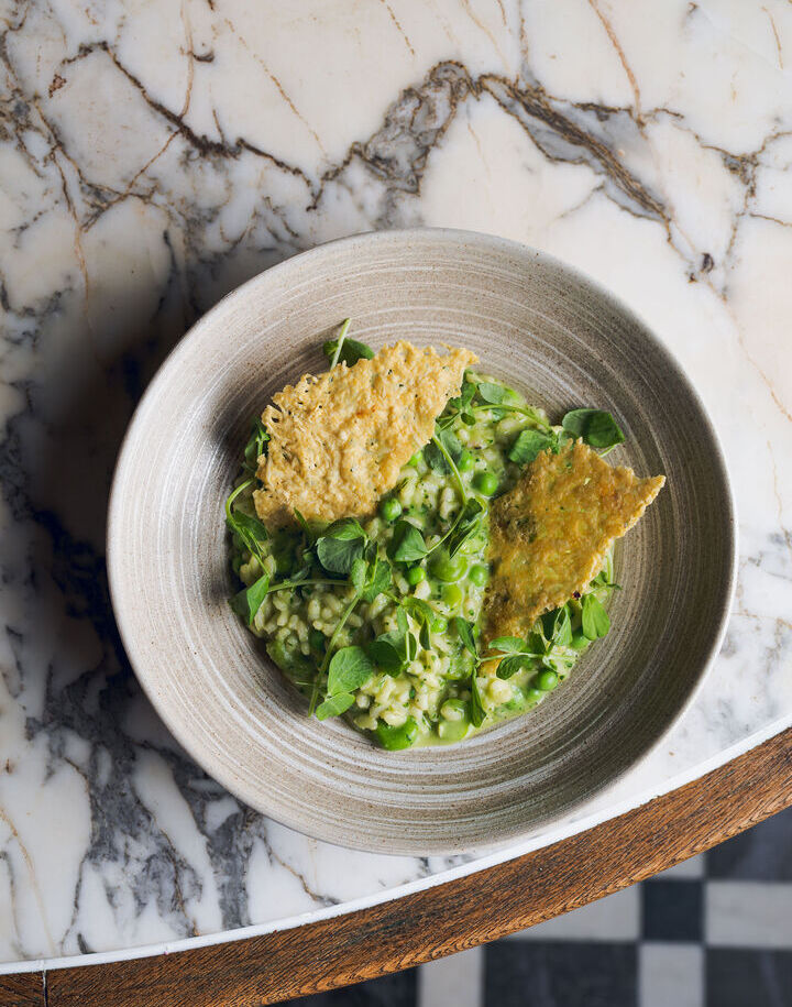 Creamy pea risotto with fresh peas, microgreens and two crisp Parmesan tuile in a rustic ceramic bowl on marble table.