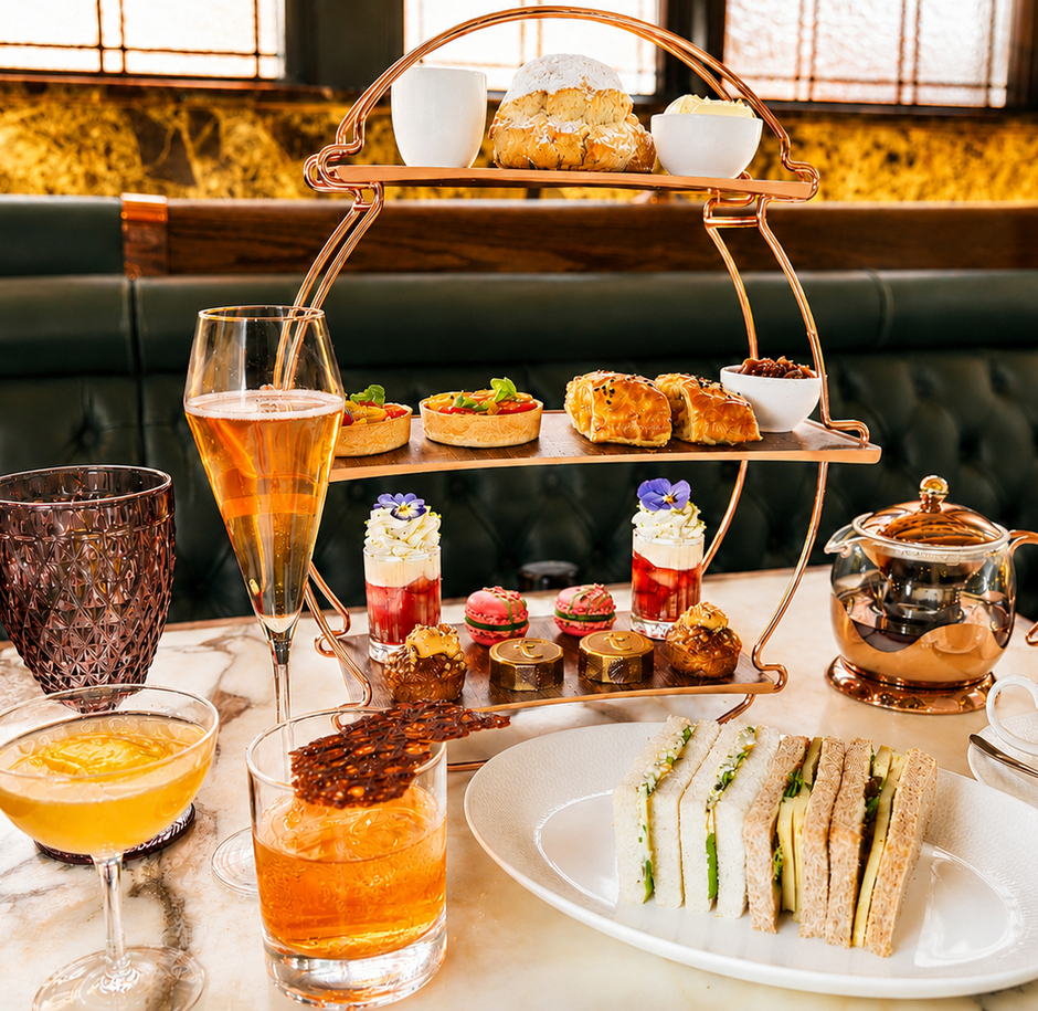 Three-tier afternoon tea stand with assorted pastries, sandwiches, cocktails and teapot on marble table in hotel lounge.
